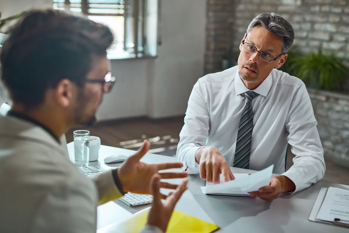 An HR interviewer in a white shirt and tie listens across a meeting table while the candidate talks with an open hand.