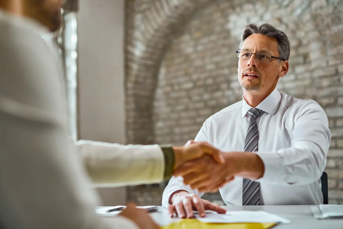 A hiring manager reaches across a desk to shake the candidate's hand at the close of an interview.