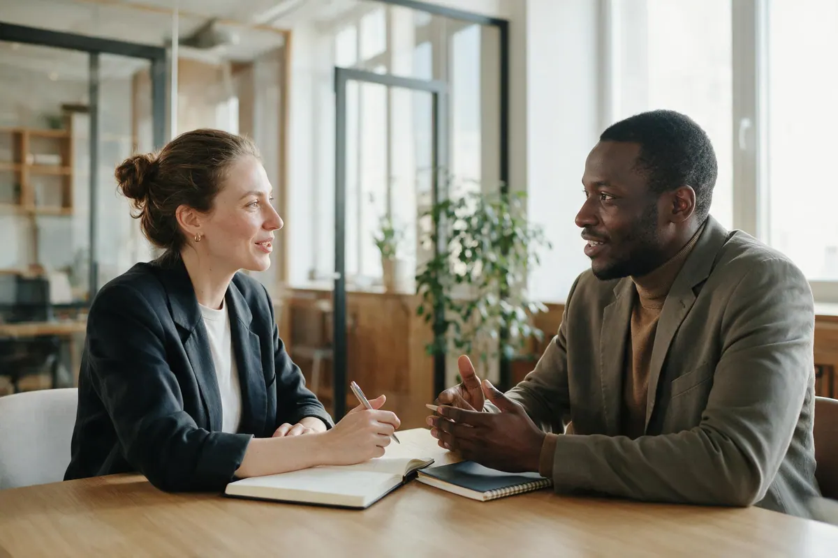 An interviewer and a candidate sit across a table in a bright office, both mid-conversation.