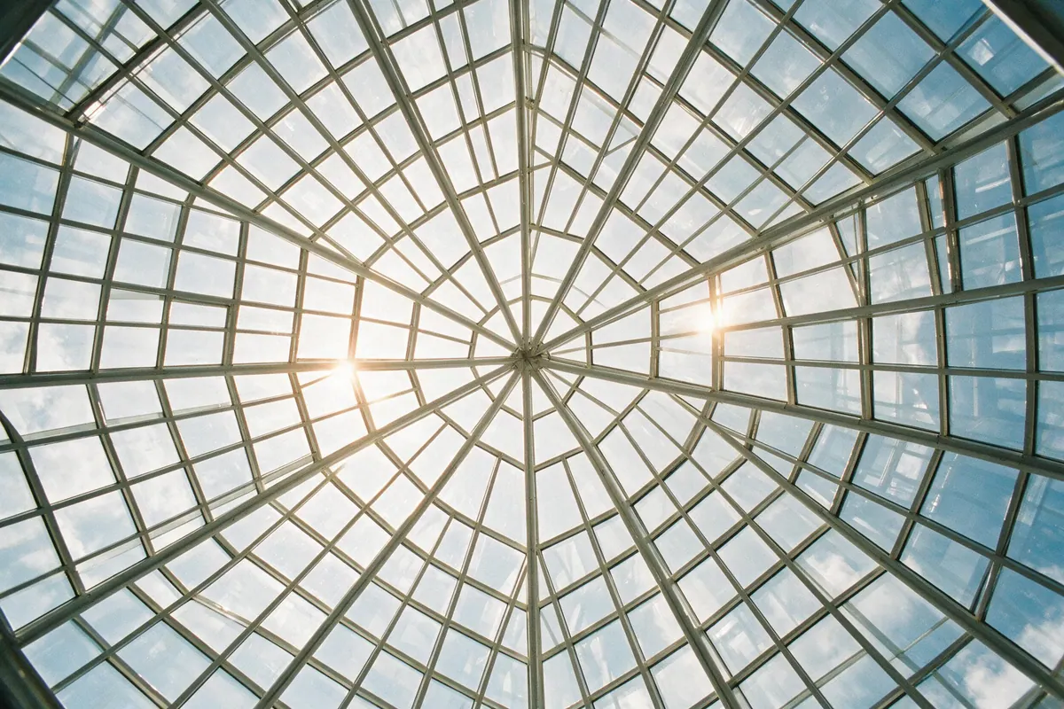 Looking straight up at a glass atrium ceiling, a steel grid framing panes of bright sky.
