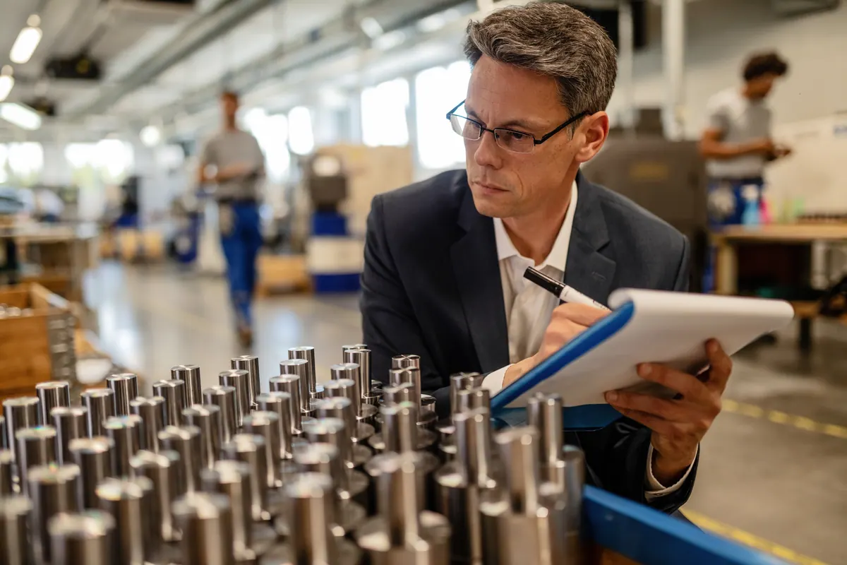 A quality inspector in safety glasses writes on a clipboard beside a rack of machined steel cylinders on a factory floor.