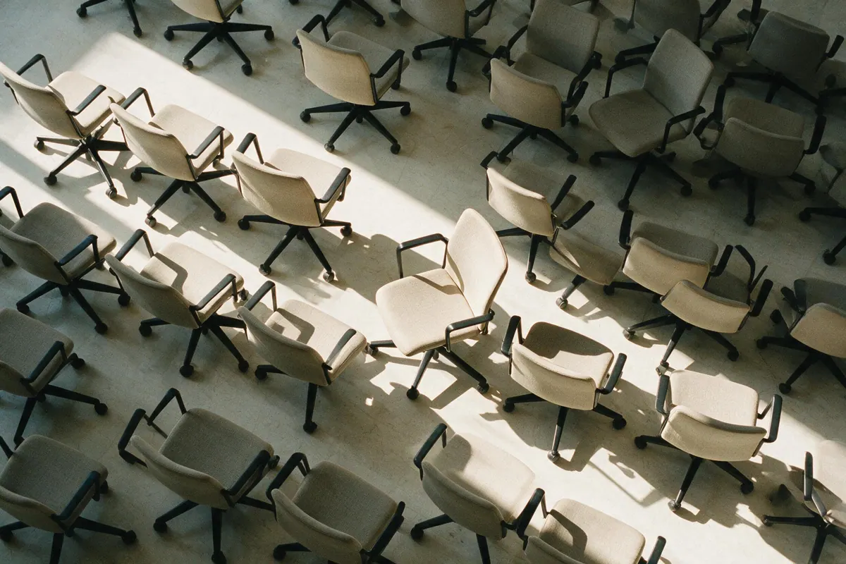 Rows of empty meeting chairs seen from directly above, sunlight cutting a diagonal across the pattern.