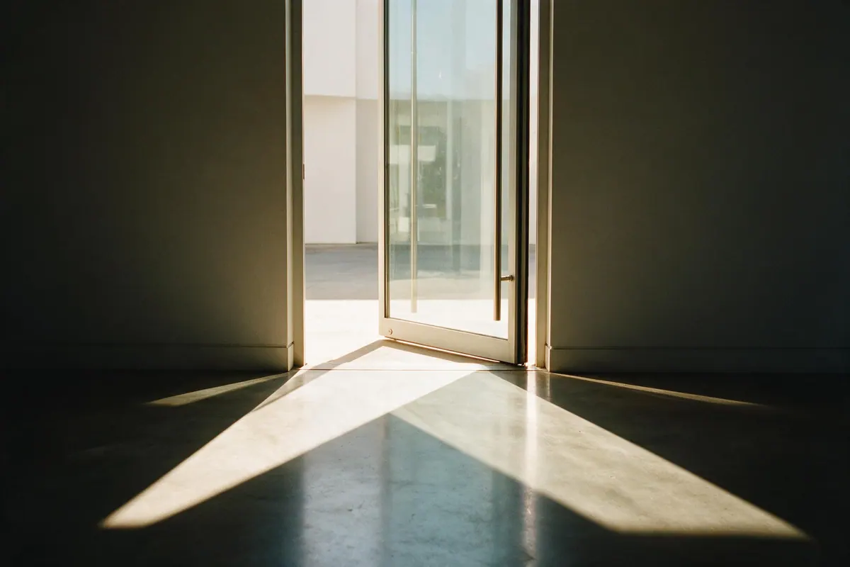 A tall glass door standing slightly ajar, daylight spilling through the gap onto a concrete floor.