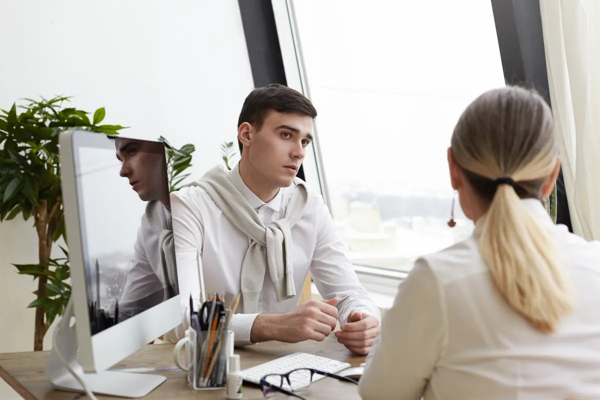 A candidate sits across a desk from an interviewer beside a bright window, listening to the next question.