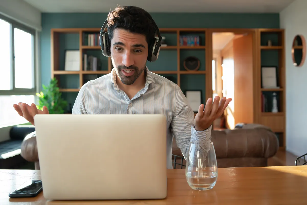 A man wearing headphones talks with both hands raised in front of an open laptop during a video interview at home.