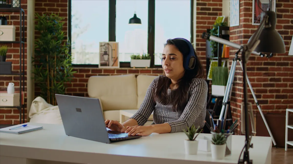 A woman in headphones sits at a laptop in a home office, mid-answer during a practice interview session.